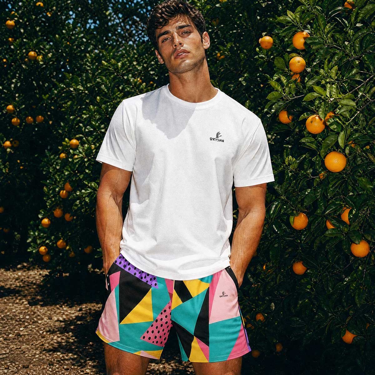 Man standing in orchard with white tee shirts and geometric colorful shorts. 