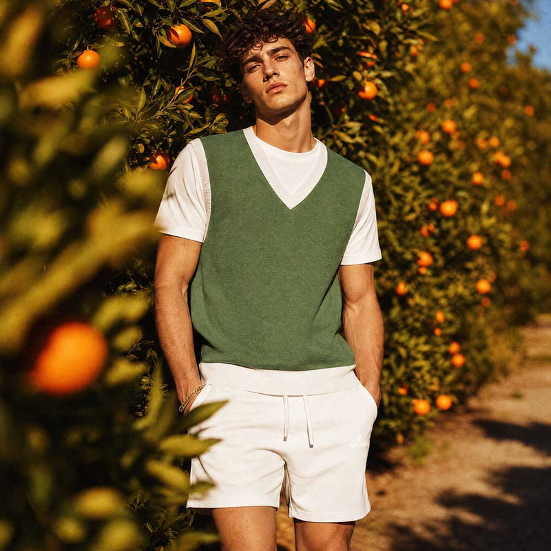 Man standing in an orange orchard wearing a green vest and white shorts.