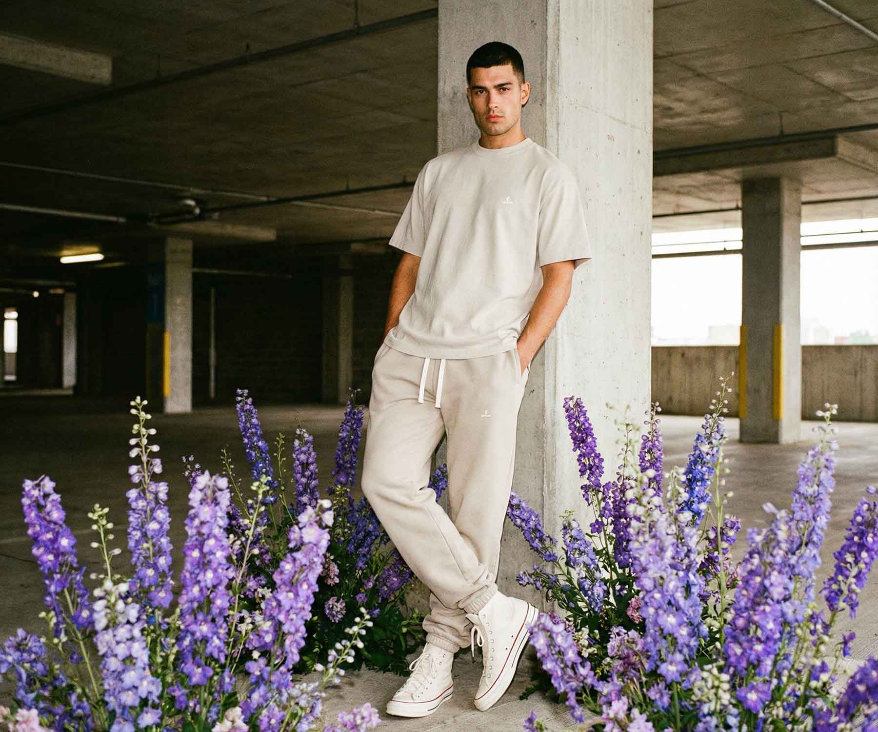 Man in beige outfit standing among purple flowers in an urban setting