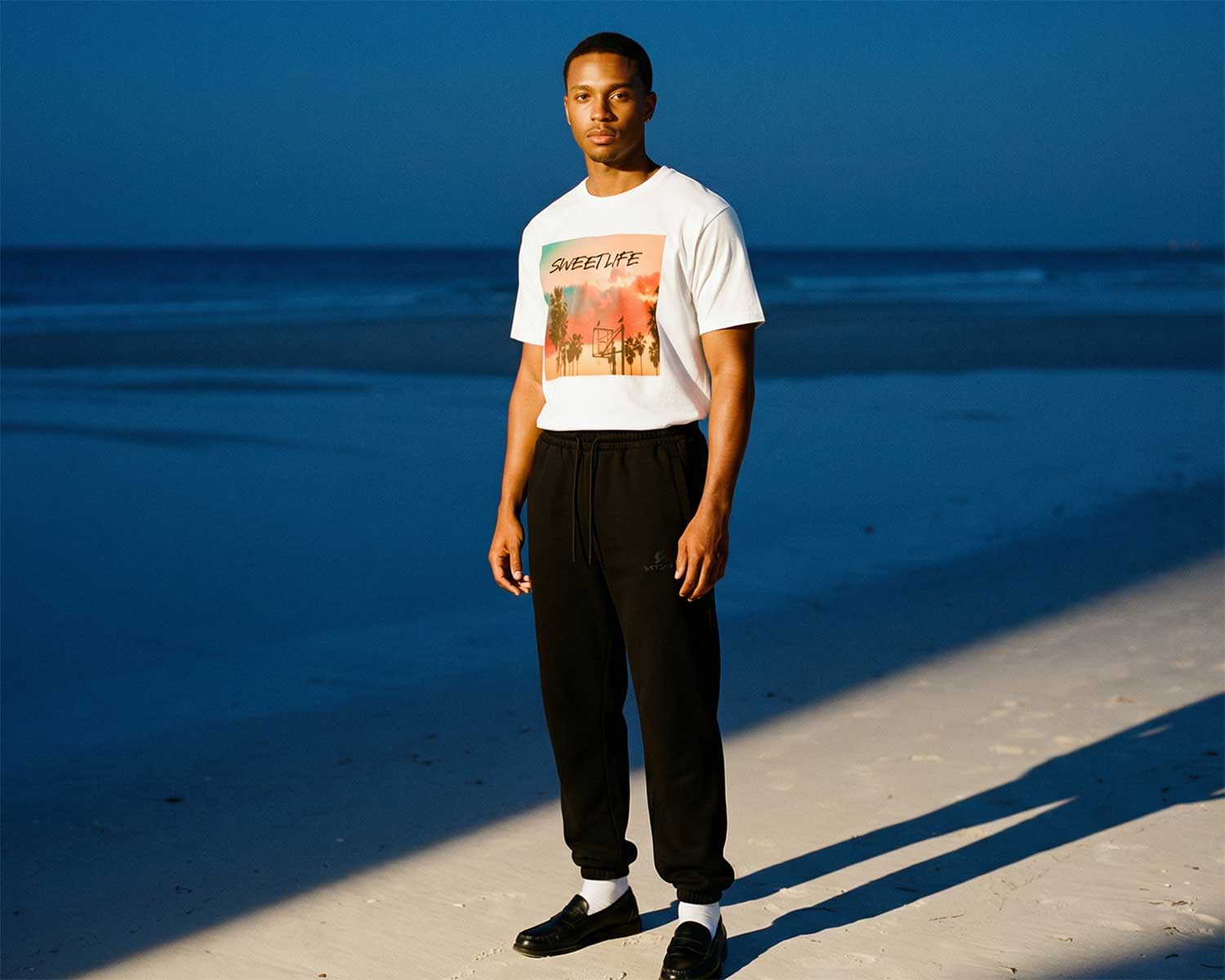 Person wearing a white t-shirt with a colorful graphic and black pants standing on a beach.