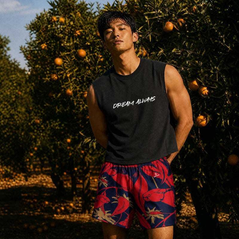 Man wearing a black sleeveless shirt with 'Dream Always' text and colorful shorts standing in an orange grove.