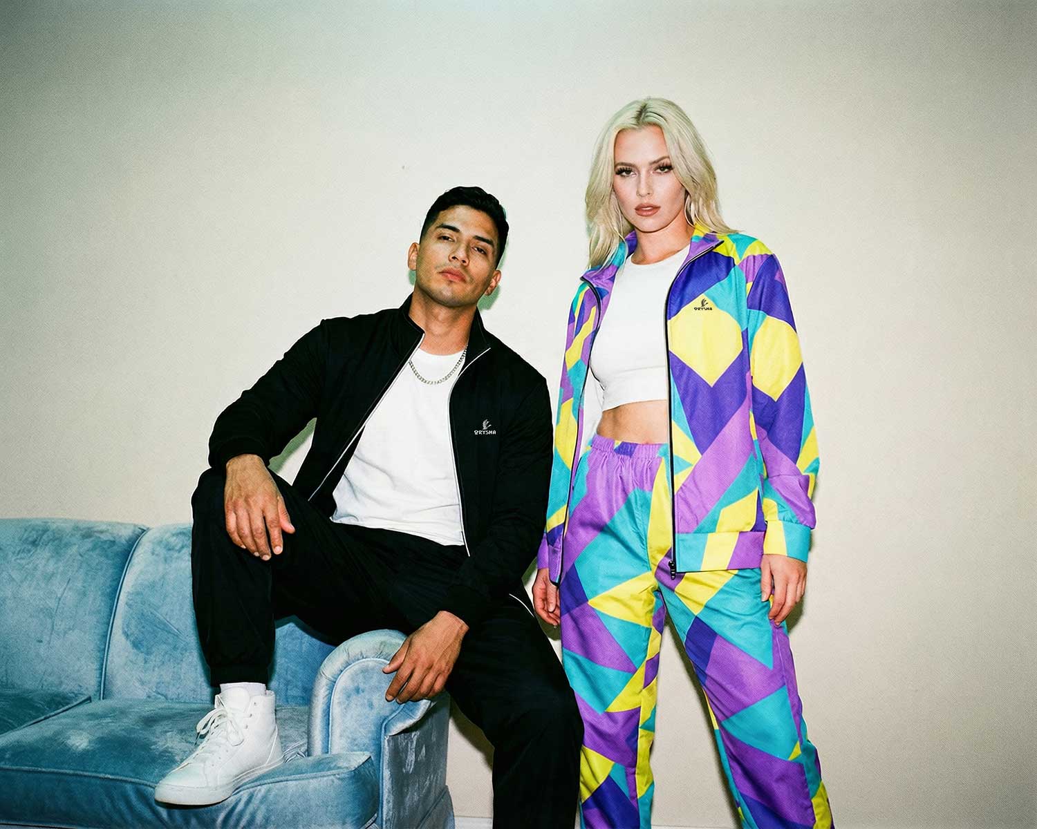 Two people sitting on a couch, one in a black outfit and the other in a colorful geometric-patterned outfit.