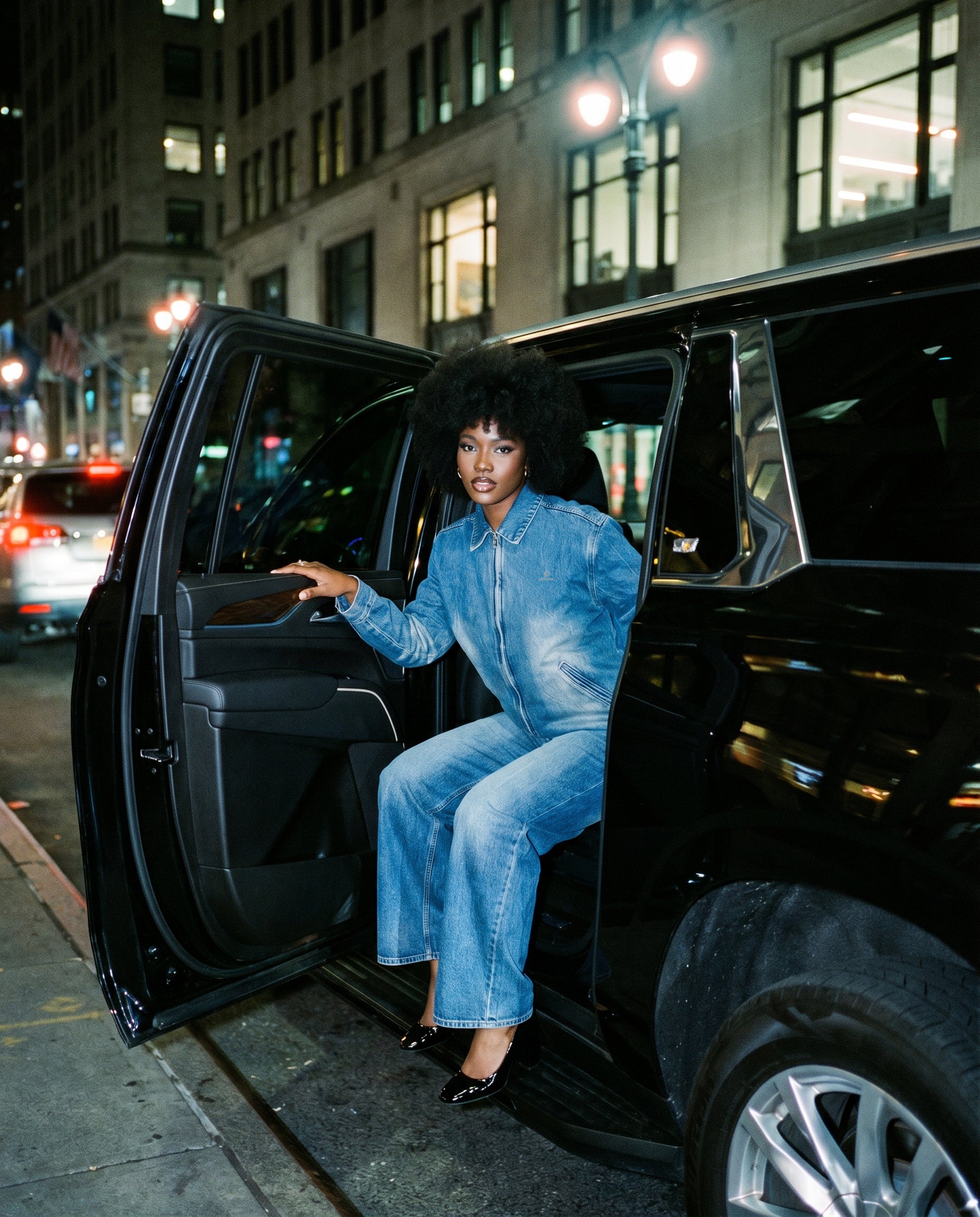 Woman in a blue outfit getting out of a black SUV on a city street at night.