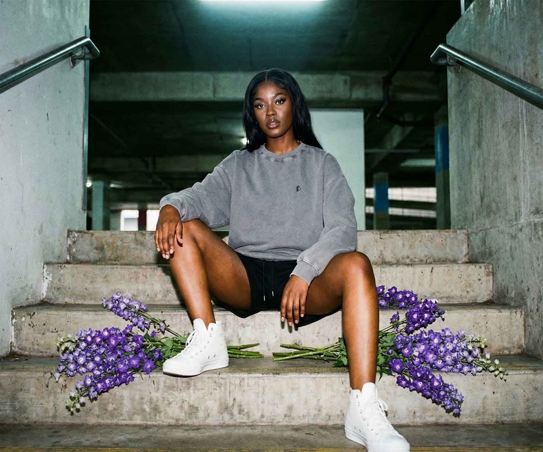 Woman sitting on concrete steps with purple flowers, wearing a gray sweatshirt and white sneakers.