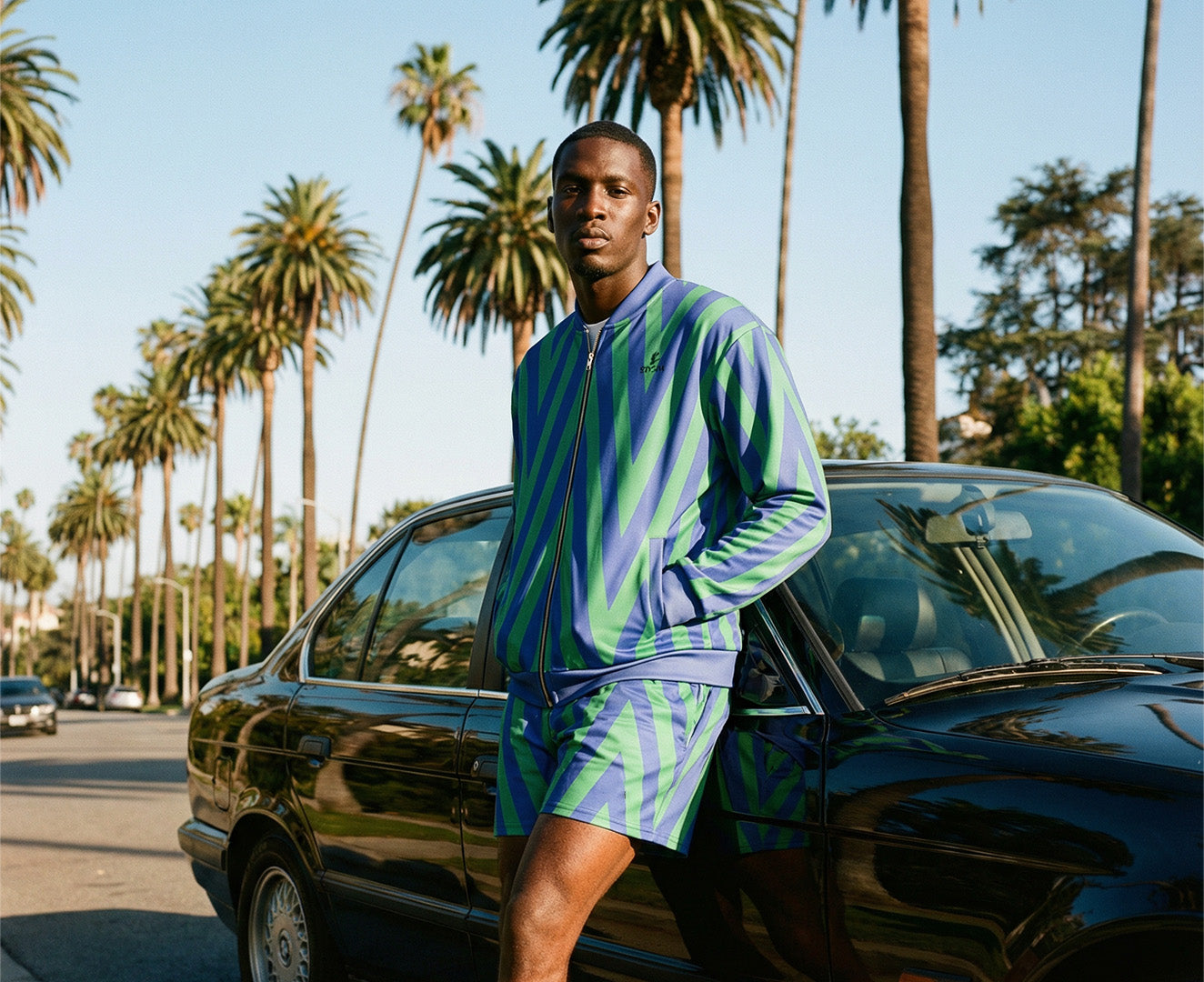 Man wearing Orysha men’s patterned track jacket and matching shorts set in blue and green geometric design, styled on a palm-lined street next to a car, modern West Coast streetwear look