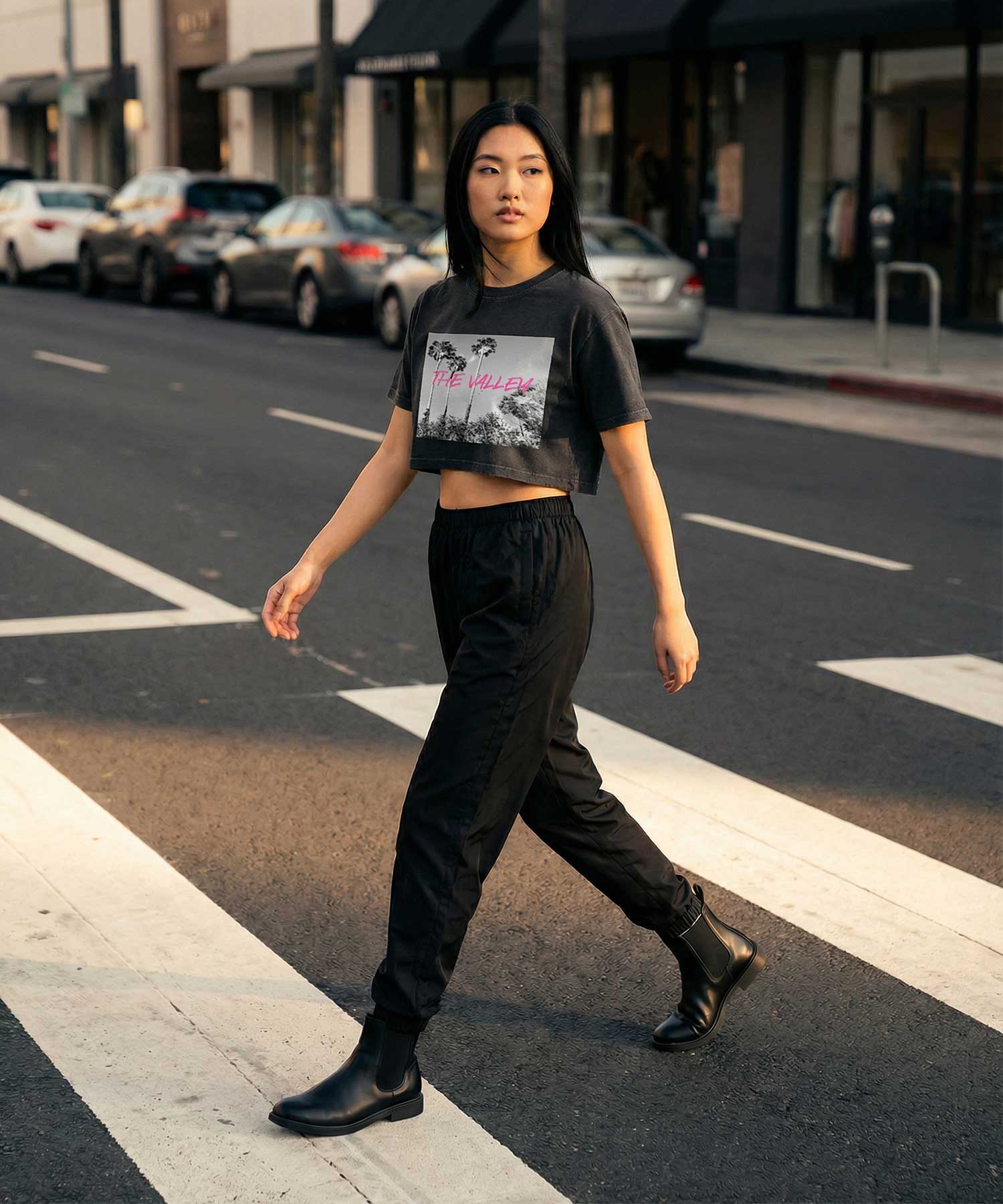 Woman wearing Orysha women’s cropped graphic t-shirt with “The Valley” palm tree design, styled with black joggers and boots while walking across a city street in an urban streetwear look
