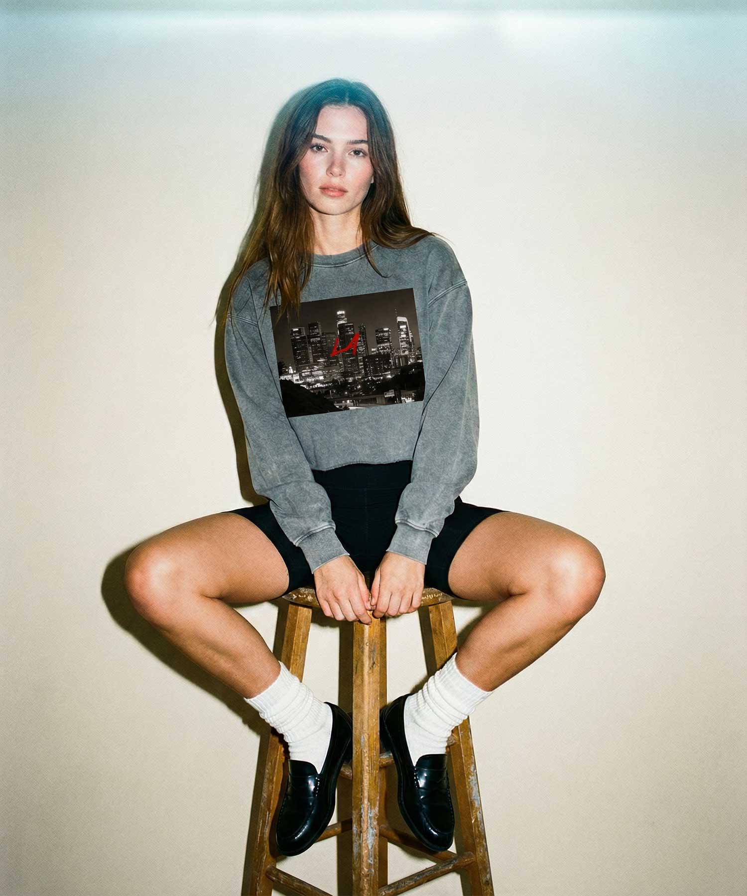 Woman wearing Orysha women’s LA skyline graphic sweatshirt with red lettering, styled with black biker shorts in a minimalist studio streetwear editorial