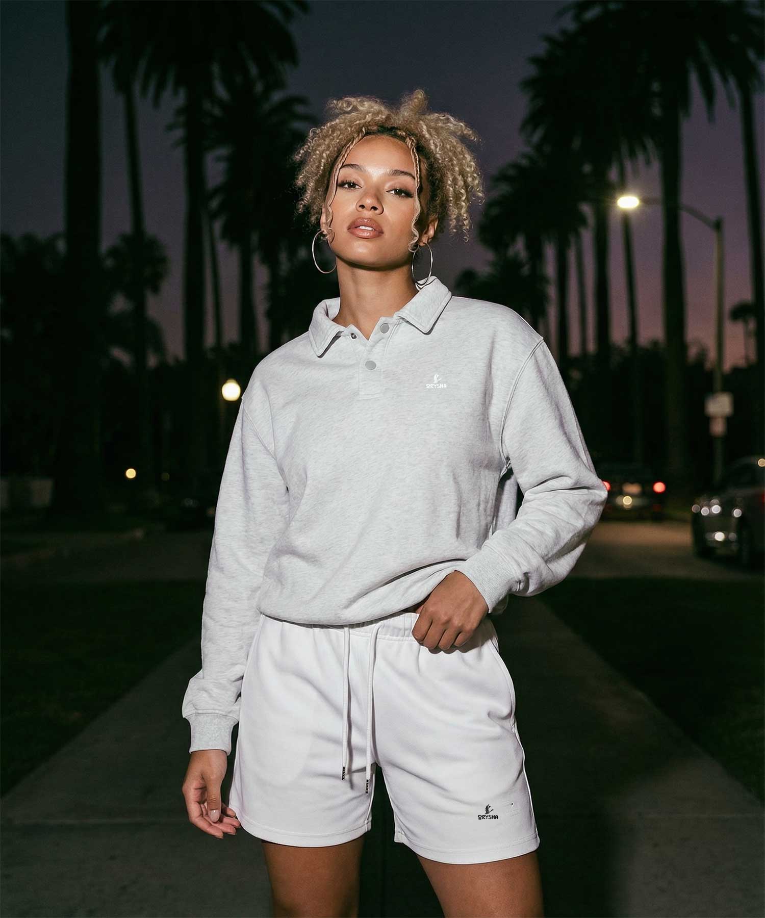 Woman wearing Orysha women’s minimalist streetwear outfit featuring a light gray polo sweatshirt and matching shorts with embroidered logo, styled on a palm-lined city street at dusk